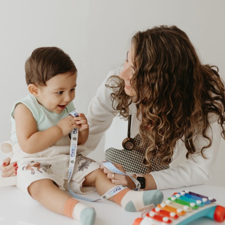 Pediatra particular Dra. Jéssica examinando bebê em consulta de puericultura com tempo e atenção Legenda: No acompanhamento de rotina, tempo e vínculo com o mesmo pediatra fazem toda a diferença.